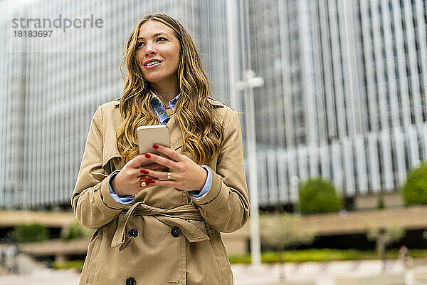 Lächelnde Geschäftsfrau hält Mobiltelefon vor Bürogebäude