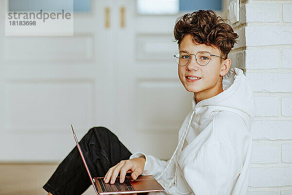 Lächelnder Junge mit Laptop  der an der Wand sitzt
