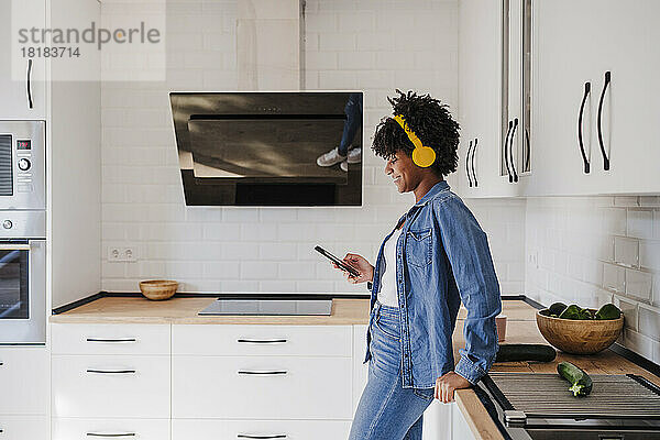 Lächelnde junge Frau mit kabellosen Kopfhörern steht zu Hause an der Küchentheke