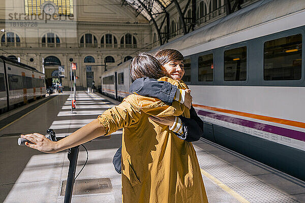 Glückliche Frau mit Tretroller umarmt Freundin am Bahnhof