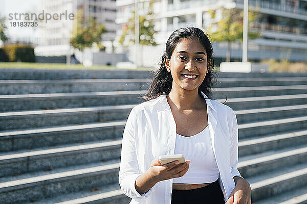 Lächelnde junge Frau mit Smartphone vor der Treppe
