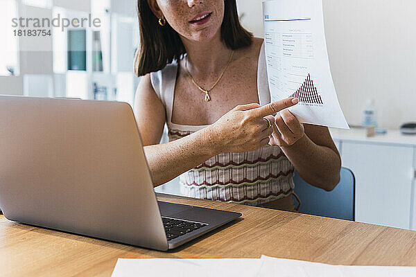 Geschäftsfrau bei Videoanruf über Laptop zeigt Diagramm im Büro