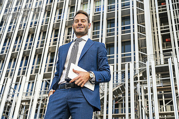 Lächelnder Geschäftsmann mit Tablet-PC steht vor Bürogebäude