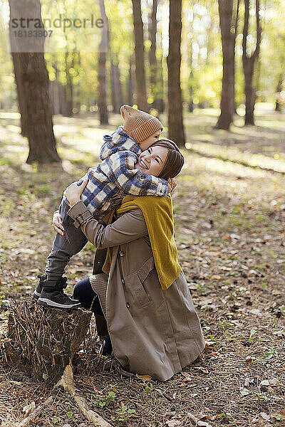 Junge steht auf Baumstumpf und umarmt Mutter im Park