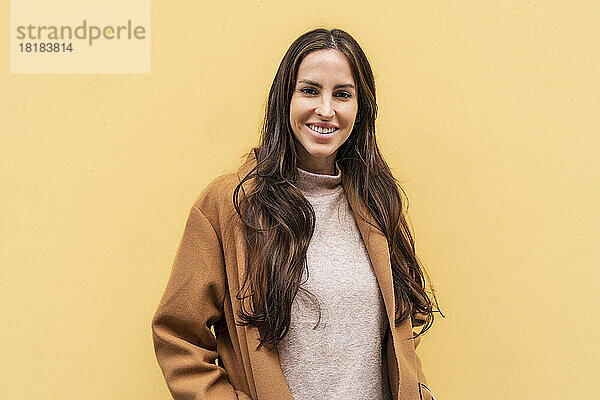 Lächelnde junge Frau mit langen Haaren vor gelber Wand