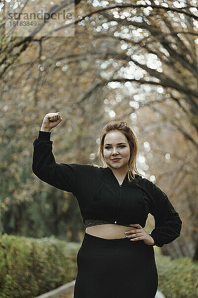 Smiling woman flexing muscles standing in autumn park