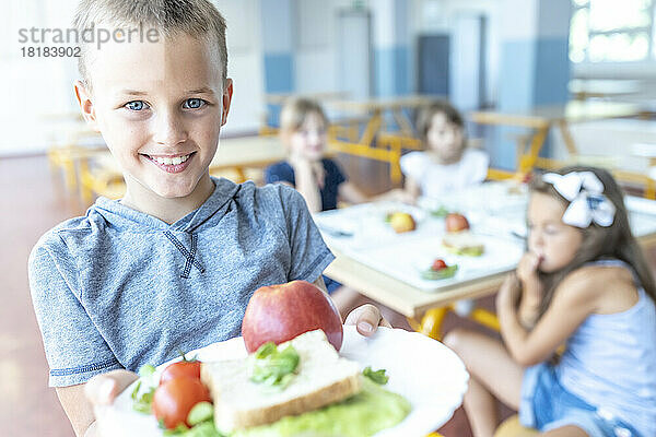 Lächelnder Junge hält gesundes Mittagessen auf dem Teller und steht in der Cafeteria
