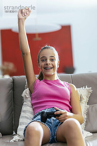 Fröhliches Mädchen mit erhobenem Arm sitzt mit Joystick zu Hause auf dem Sofa