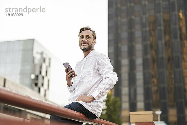 Lächelnder Mann sitzt mit Smartphone am Geländer