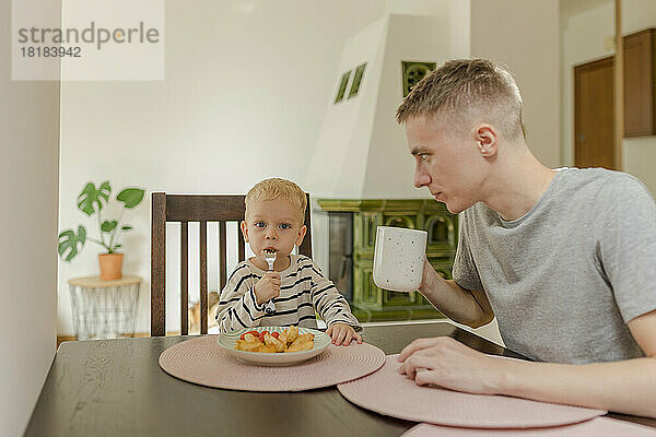 Netter Junge isst Essen  während sein Vater eine Teetasse am Esstisch hält