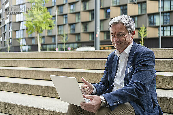 Lächelnder reifer Geschäftsmann gestikuliert bei Videoanruf über Laptop auf Schritte