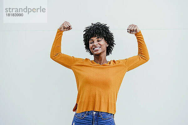 Lächelnde Afro-Frau gestikuliert Stärke vor weißer Wand