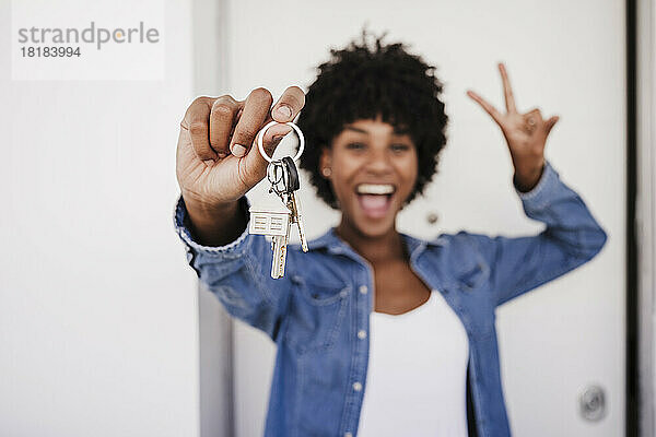 Fröhliche Frau zeigt Hausschlüssel und Peace-Zeichen vor der Tür