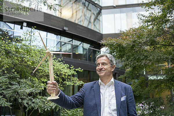 Lächelnder reifer Geschäftsmann mit Windturbinenmodell  der im Büropark steht