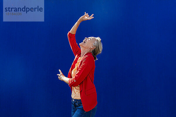 Fröhliche Frau im roten Blazer tanzt an der blauen Wand