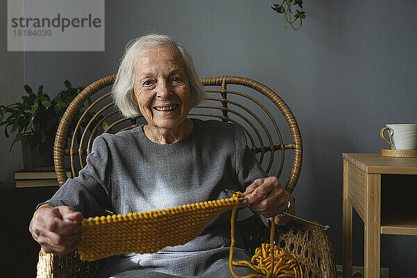 Lächelnde ältere Frau mit Strickwolle und Nadel auf dem Stuhl zu Hause