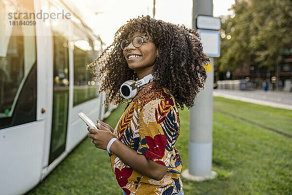Glückliche junge Frau steht mit Smartphone an der Straßenbahn