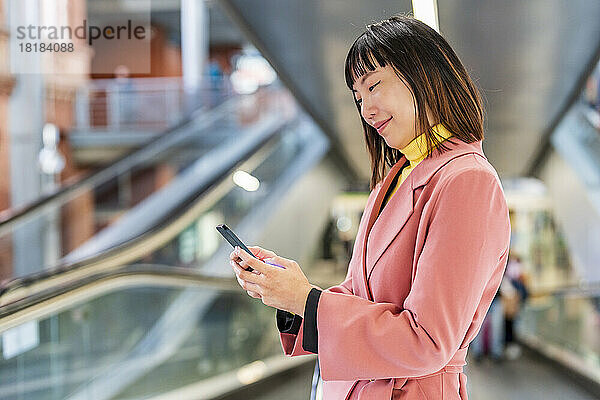 Lächelnde junge Frau  die auf der Rolltreppe Textnachrichten über ihr Smartphone sendet