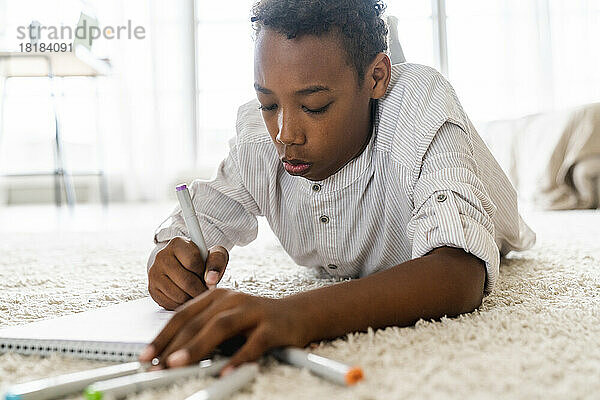 Junge schreibt mit Filzstift in ein Buch  das auf dem Teppich liegt