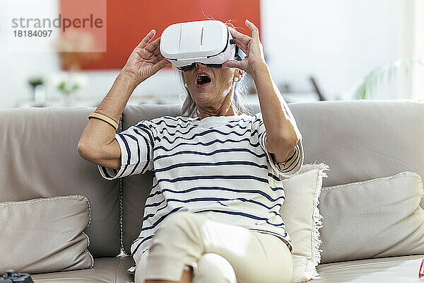 Senior woman with mouth open wearing virtual reality headset sitting on sofa at home