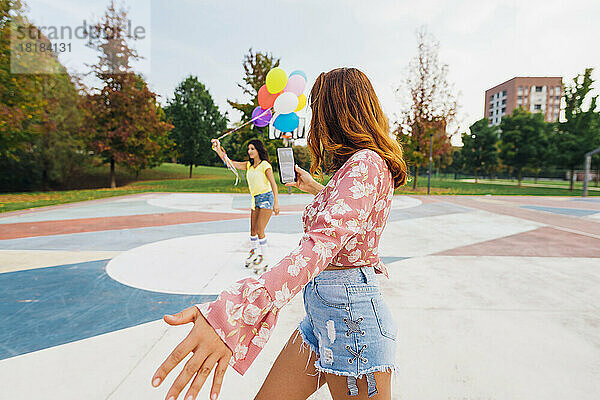 Frau fotografiert Freundin mit Luftballons und Rollschuhlaufen per Handy auf Sportplatz