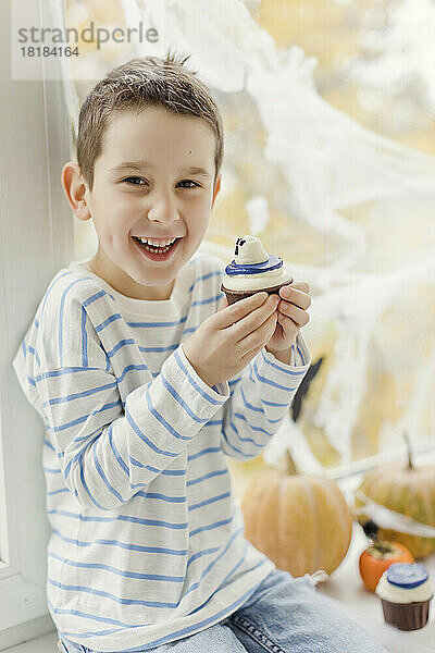 Glücklicher Junge mit Cupcake  der zu Hause vor dem Fenster sitzt