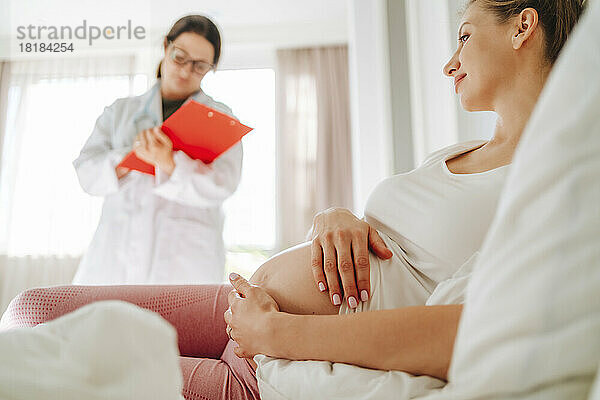Lächelnde schwangere Frau liegt auf dem Bett  mit dem Arzt im Hintergrund