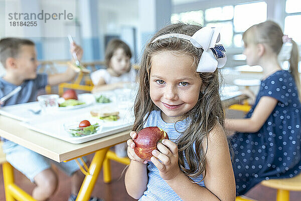 Lächelndes Mädchen hält Apfel in der Hand und sitzt mit Freunden in der Schulcafeteria