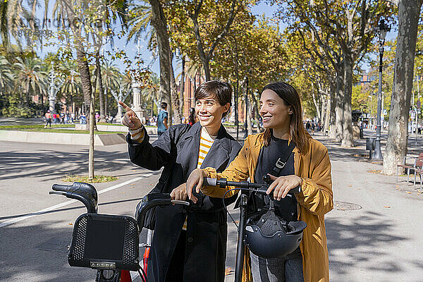 Lächelnde Freunde mit Elektrofahrrad und Tretroller diskutieren auf Fußweg
