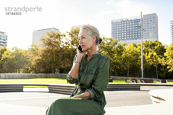 Nachdenkliche reife Frau  die auf einer Bank sitzt und über ihr Smartphone spricht