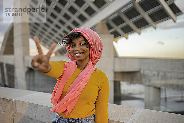 Lächelnde Frau mit Kopftuch zeigt eine Geste des Friedenszeichens und steht an der Wand