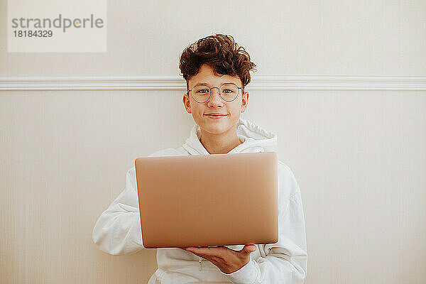 Happy boy sitting with laptop in front of wall