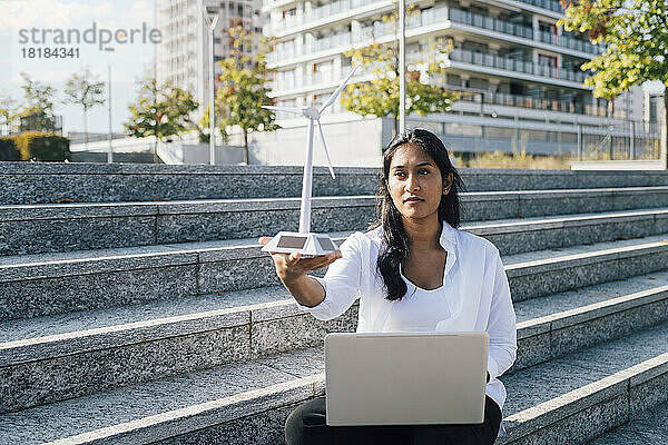 Junge Frau mit Windturbinenmodell und Laptop sitzt auf Stufen