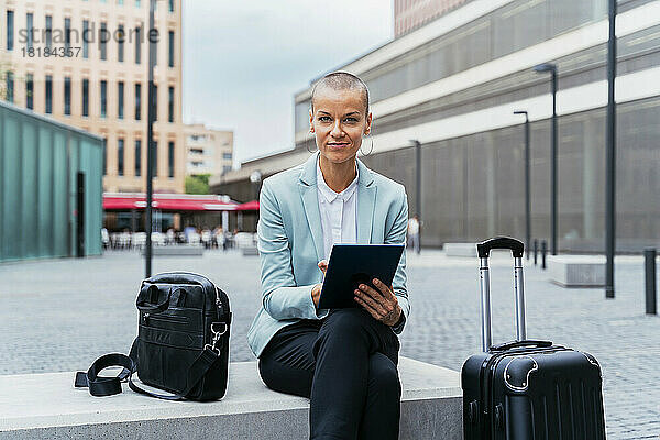 Lächelnde Geschäftsfrau mit Tablet-PC und Gepäck sitzt auf Bank am Fußweg