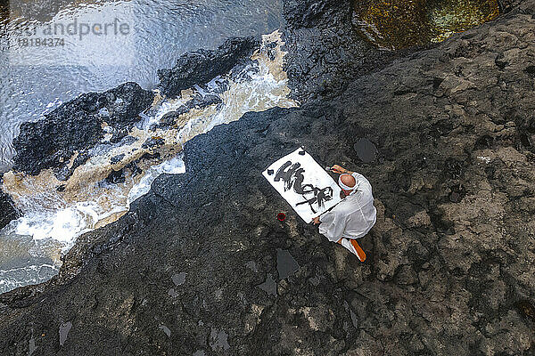 Reifer Mönch macht Kalligraphie und sitzt auf einem Felsen am Meer