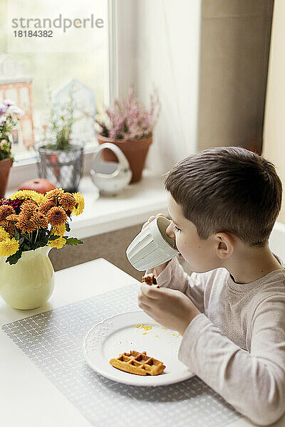 Junge trinkt heiße Schokolade aus einer Tasse auf dem Esstisch in der Küche
