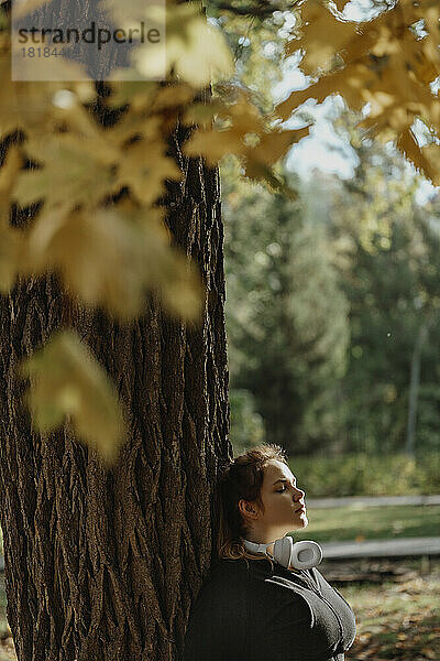 Junge Frau mit geschlossenen Augen lehnt an Baumstamm im Park