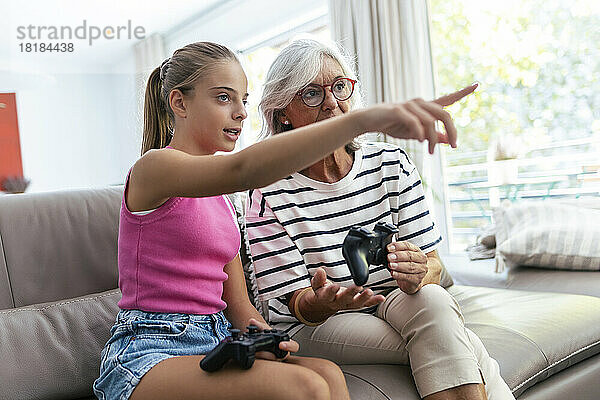 Enkelin zeigt auf Großmutter  die zu Hause mit Joystick auf dem Sofa sitzt