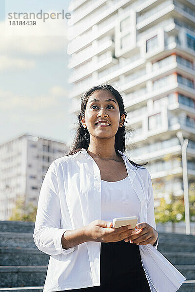 Lächelnde junge Frau mit Mobiltelefon steht vor dem Gebäude