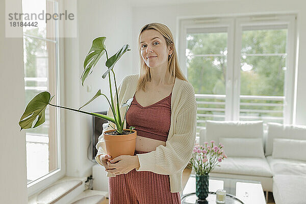 Lächelnde junge Frau mit Monstera-Pflanze steht zu Hause im Wohnzimmer