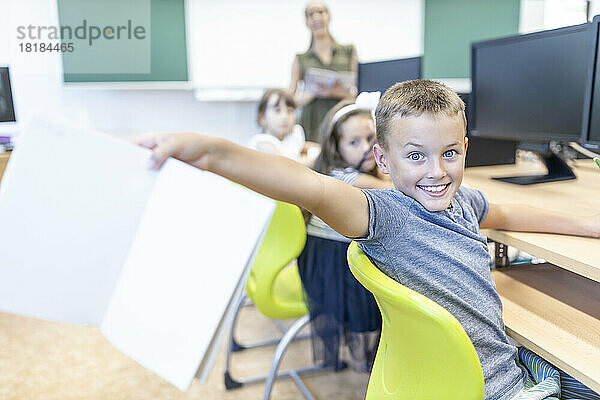 Aufgeregter Junge zeigt Notizbuch auf Stuhl im Klassenzimmer sitzend