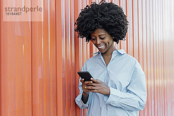 Lächelnde junge Frau benutzt Smartphone und steht an orangefarbener Wand