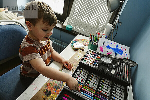 Lächelnder Junge wählt Buntstiftfarbe aus der Schachtel auf dem heimischen Tisch