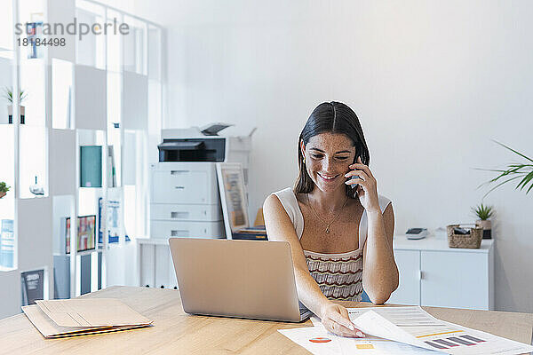 Lächelnde Geschäftsfrau  die im Büro mit dem Mobiltelefon spricht und Dokumente abholt