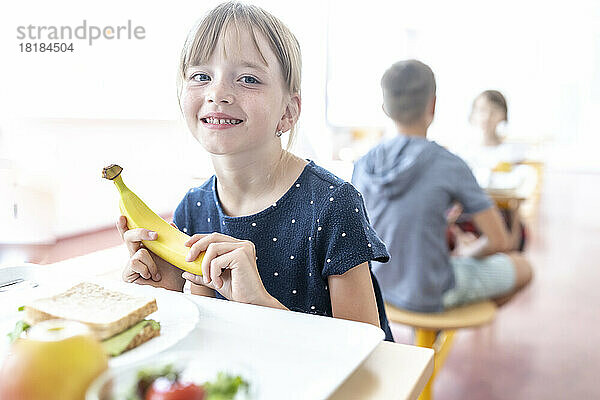 Smiling student holding fresh banana at lunch break in cafeteria
