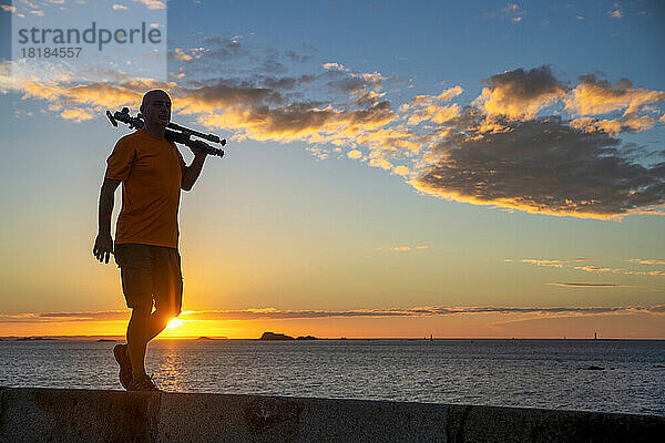 Reifer Mann läuft bei Sonnenuntergang mit Stativ auf dem Pier