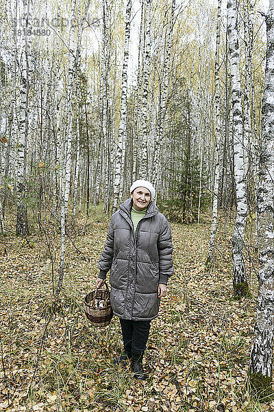 Lächelnde ältere Frau hält einen Korb voller Pilze im Wald