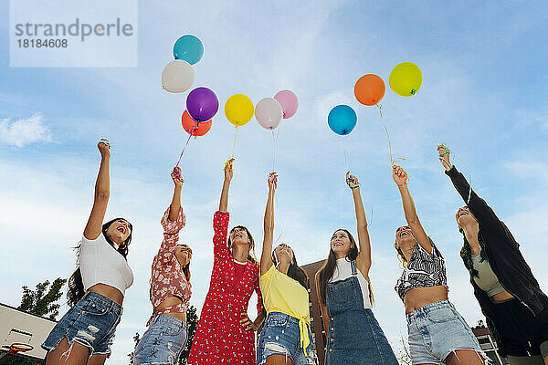 Fröhliche Freunde mit erhobenen Händen und bunten Luftballons unter blauem Himmel