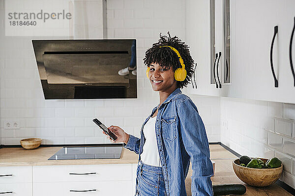 Glückliche Frau mit kabellosen Kopfhörern  die zu Hause mit ihrem Smartphone steht