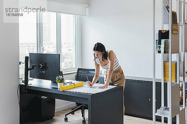 Junger Architekt lehnt am Schreibtisch und analysiert den Bauplan im Büro